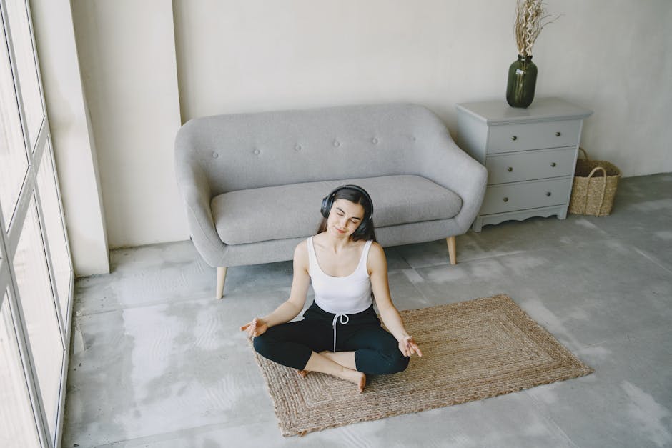 A young woman sits on a mat in a contemporary living room, meditating with headphones on.