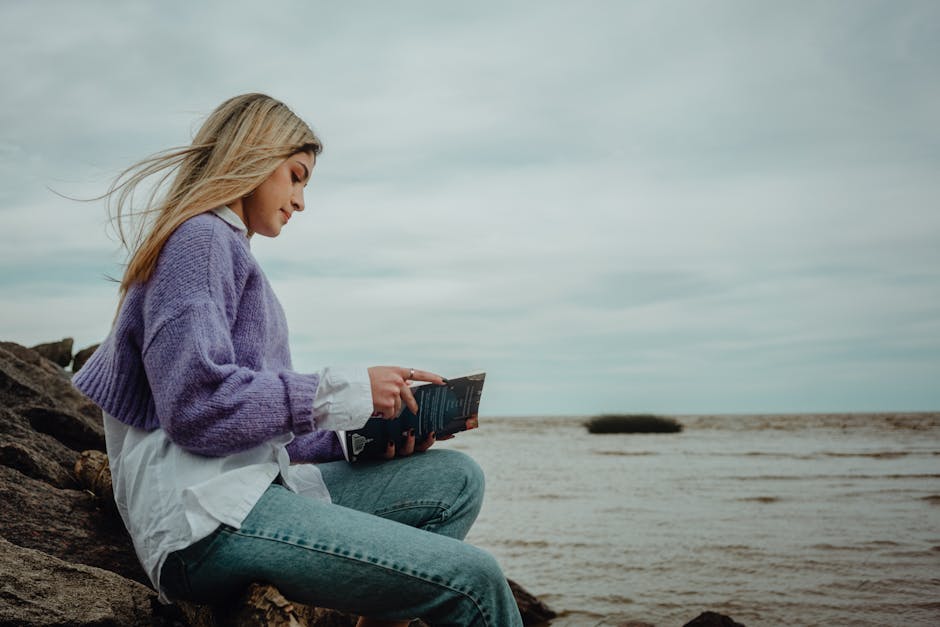 A woman in a purple sweater reads a book by the seaside, exuding serenity and relaxation.