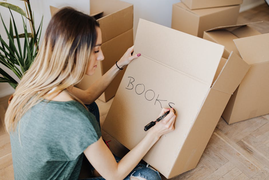 From above side view glad young lady in casual shirt writing on carton box word books while packing stuff and preparing to move in new apartment