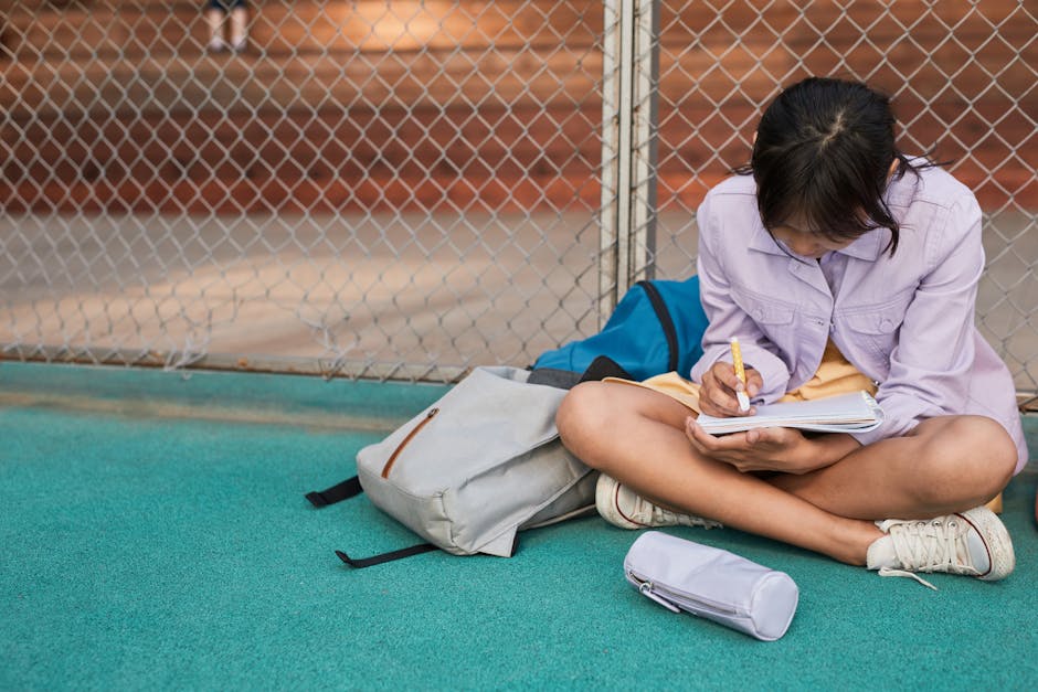 Teen girl sitting outdoors writing in a notebook by a fence.