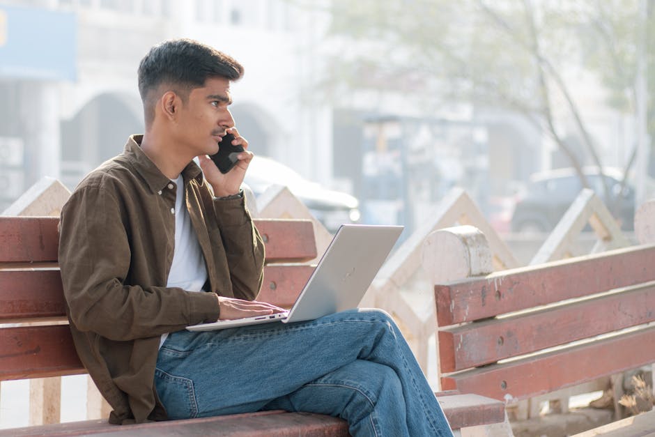 A young man multitasking with a laptop and smartphone on a park bench, embracing remote work outdoors.